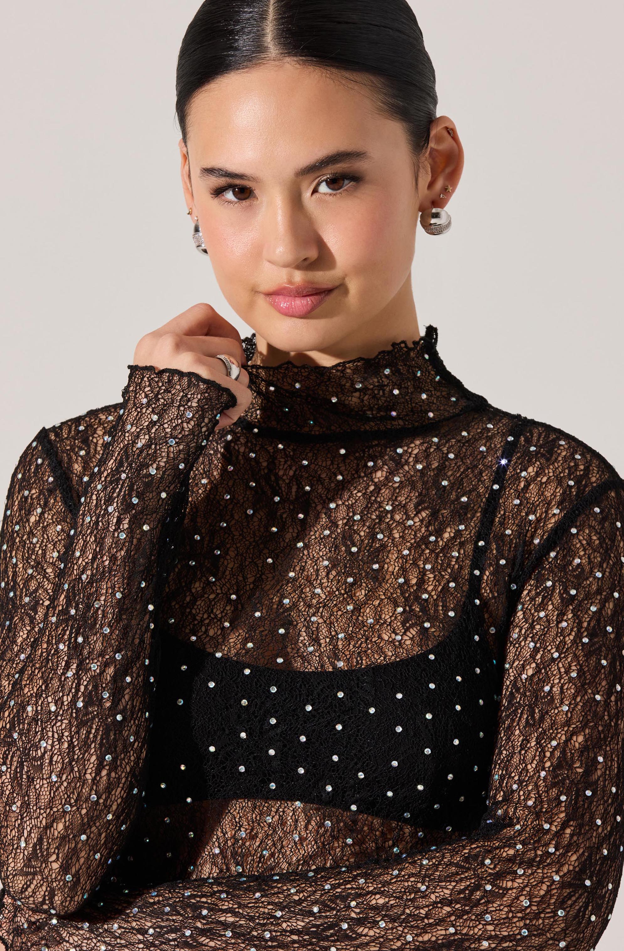 The woman is wearing a sheer black lace top with long sleeves and a high neckline, adorned with small, evenly spaced rhinestones. Underneath, she has a black camisole with thin straps and a pattern of rhinestones that complement the lace top. She is accessorized with silver earrings and a ring.