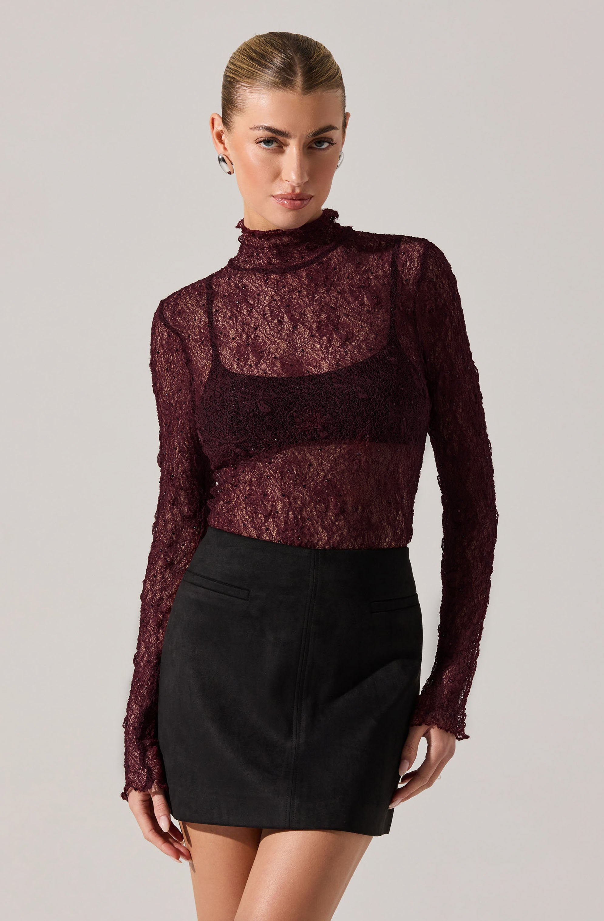 A woman is wearing a sheer, long-sleeved, burgundy lace turtleneck top paired with a black high-waisted skirt. The lace top features intricate floral patterns and a ruffled trim at the neckline.