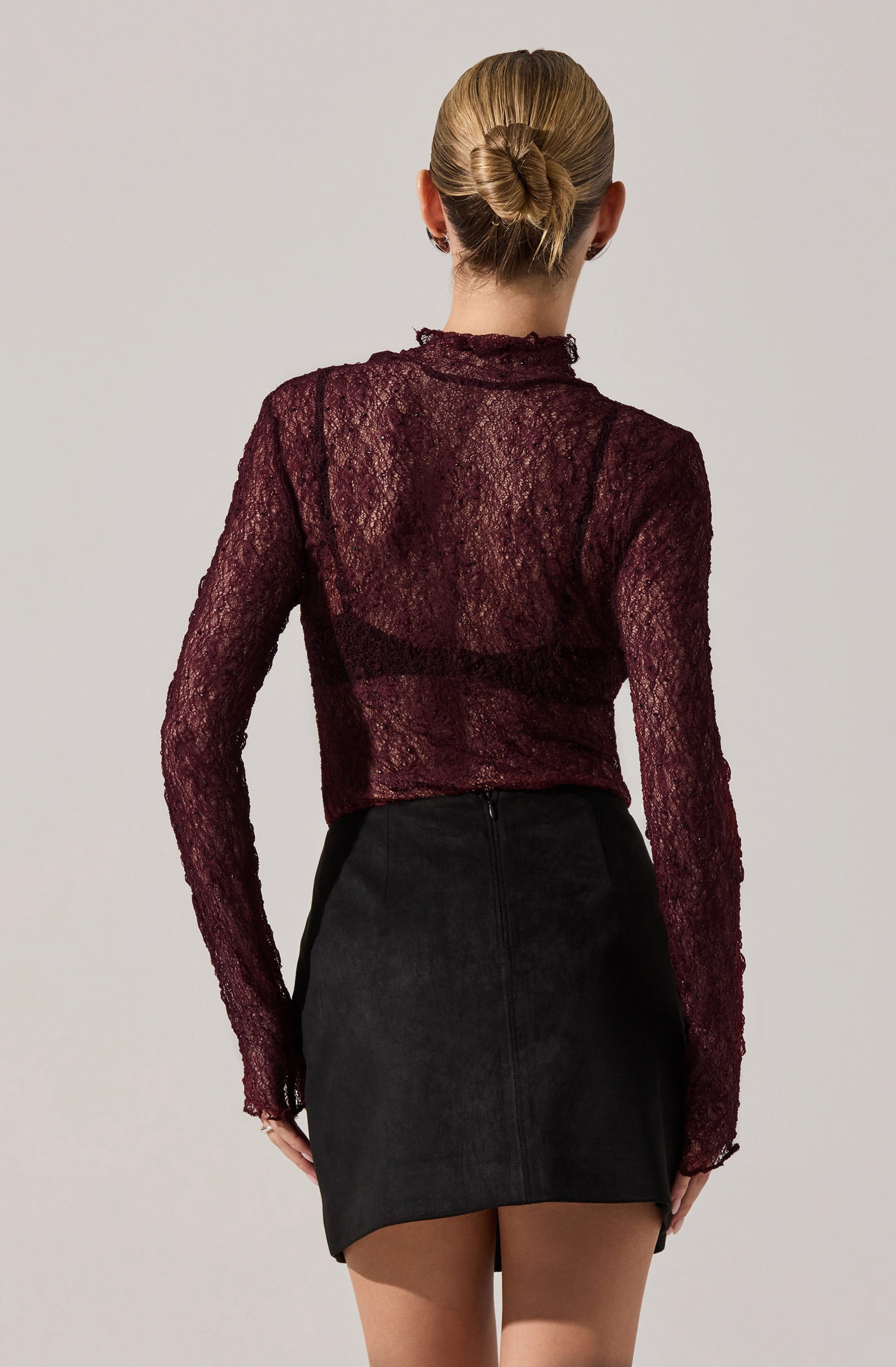 A person is wearing a semi-sheer, long-sleeved, dark red lace top with a high neckline and a slightly ruffled collar. They are also wearing a black, high-waisted, A-line mini skirt made of a textured fabric, possibly suede.