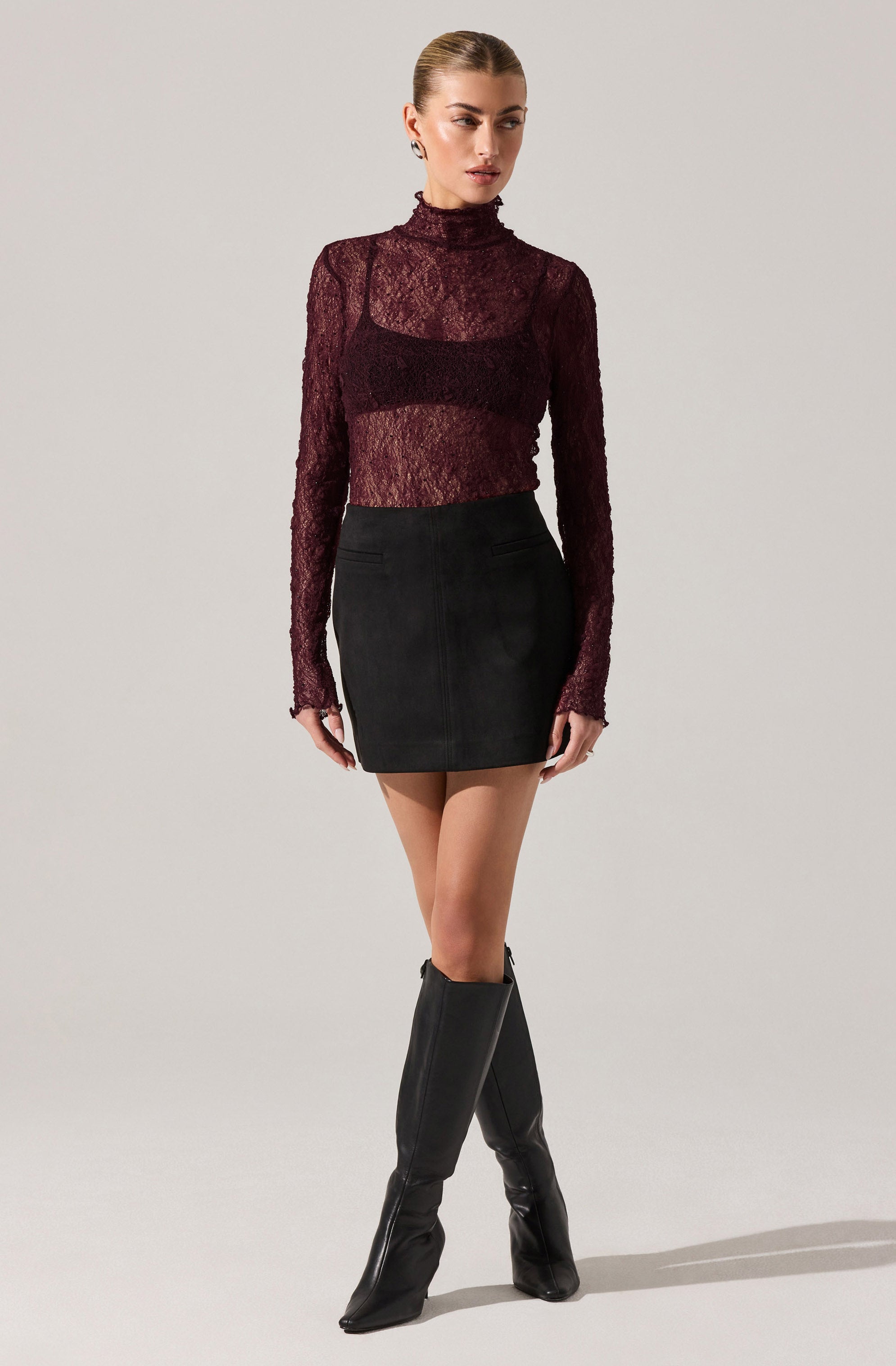 A woman is wearing a long-sleeved, burgundy lace turtleneck top that is semi-sheer, paired with a high-waisted black mini skirt. She is also wearing knee-high black boots with a slight heel.