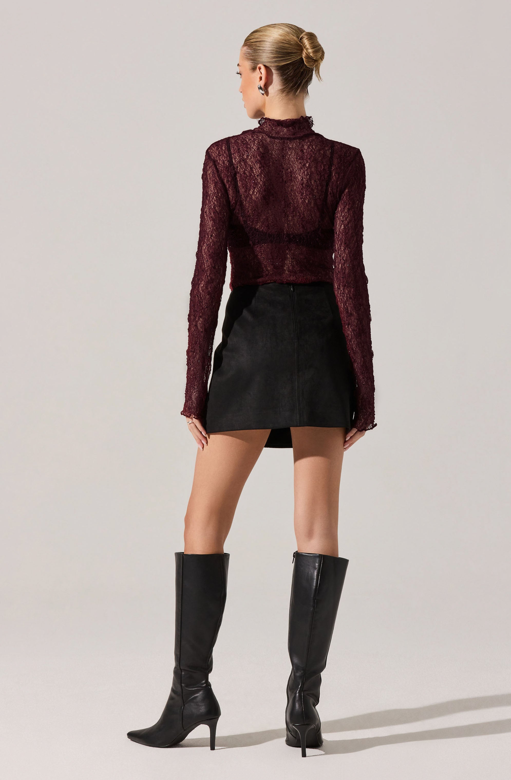 A woman is wearing a long-sleeve, semi-sheer, maroon lace top with a high neckline. She has paired it with a high-waisted, black mini skirt and black knee-high boots with pointed toes and stiletto heels.