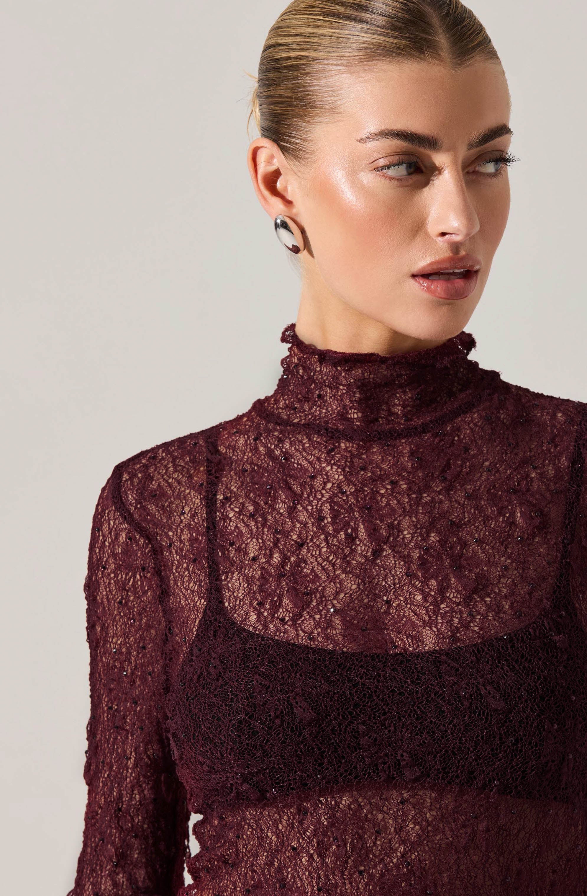A woman is wearing a high-neck, long-sleeved burgundy lace top that is sheer and textured. She has on statement earrings featuring a curved, abstract black-and-white design.