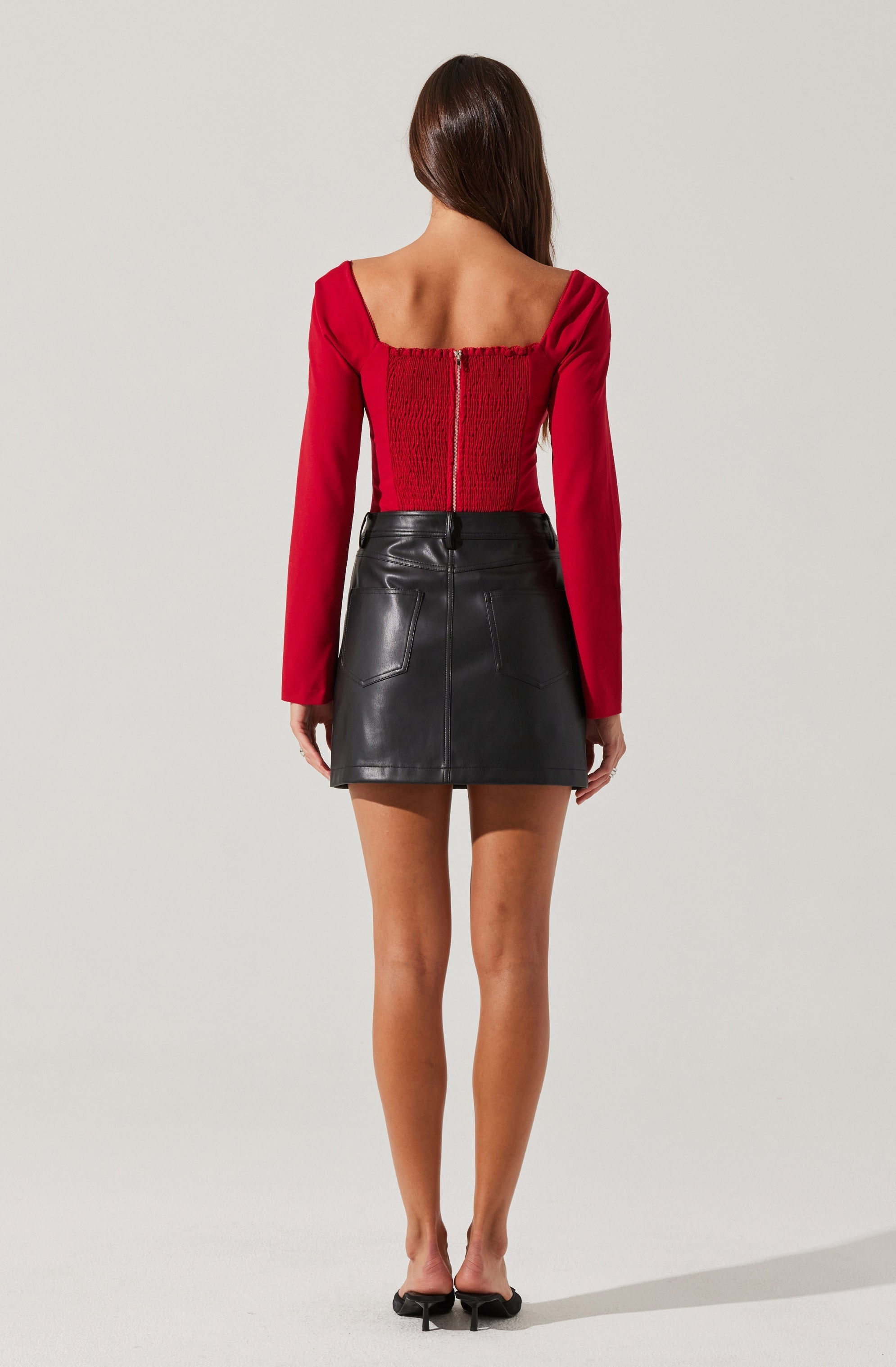 A woman is wearing a red long-sleeve top with a gathered back and a central zipper, paired with a high-waisted black leather mini skirt with two back pockets. She is also wearing black open-toe heels.