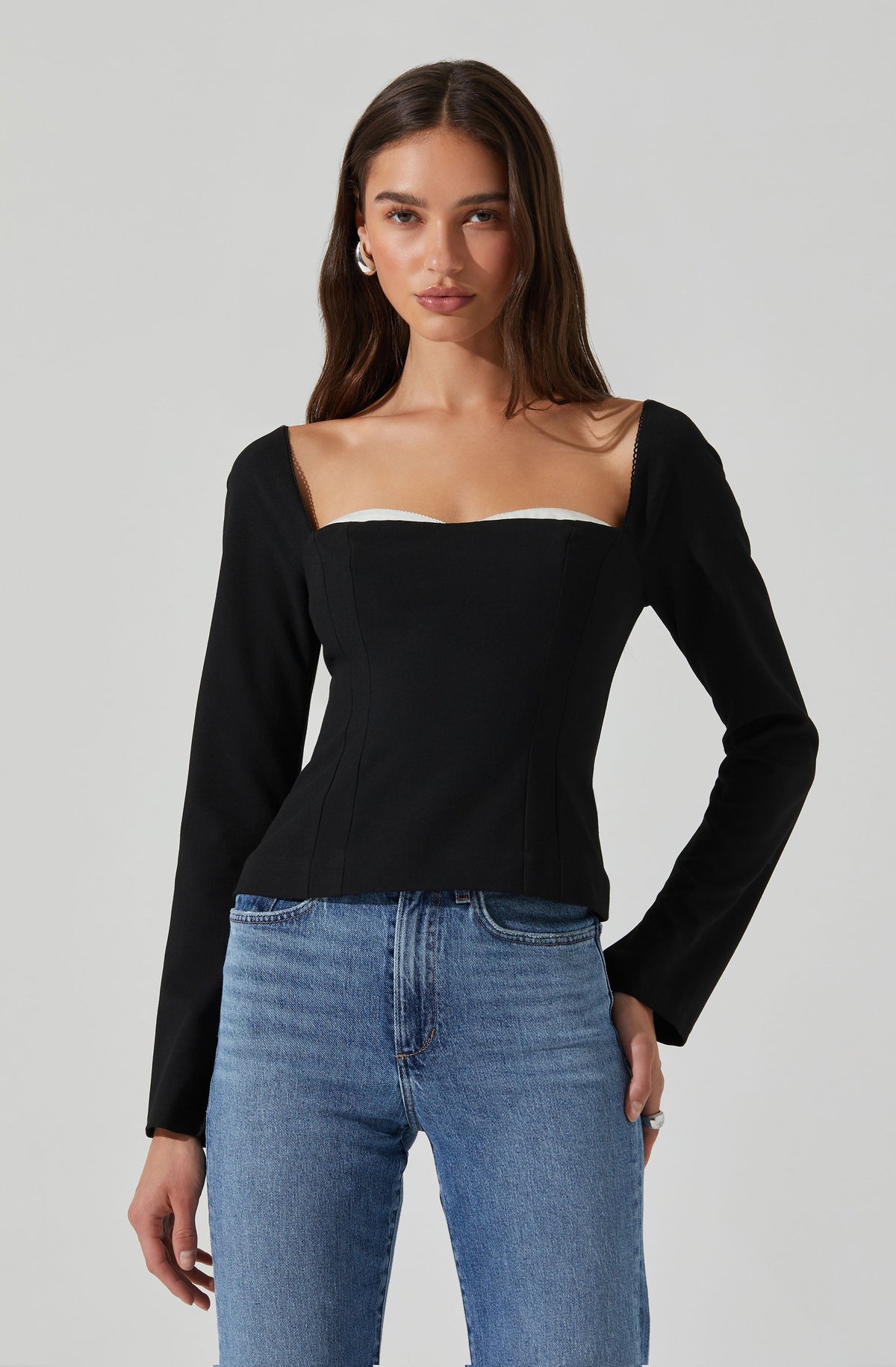 A woman is wearing a black long-sleeve top with a fitted bustier style bodice, featuring a sweetheart neckline and subtle shoulder seam details. The top is paired with light blue high-waisted jeans.