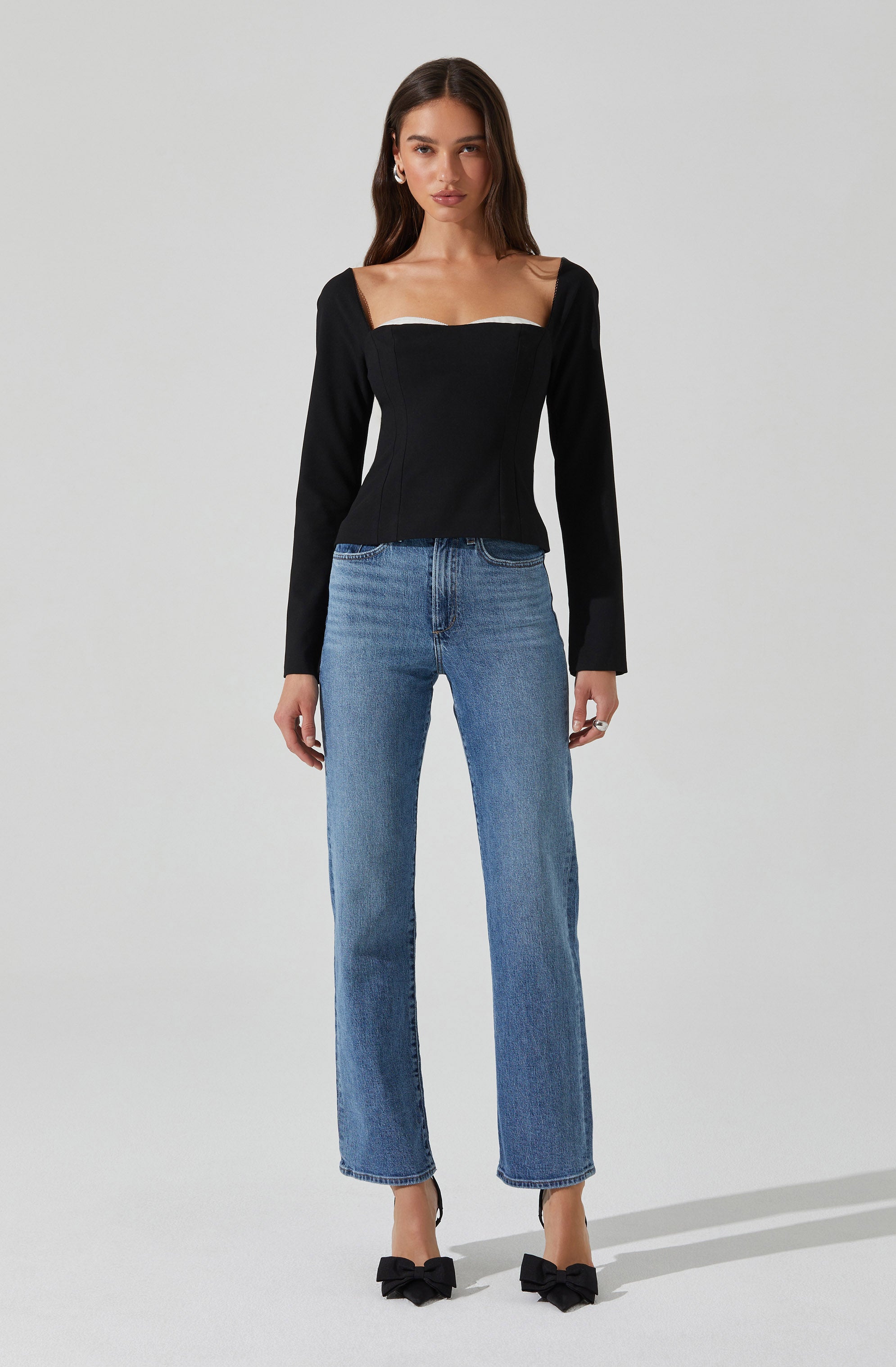 A woman is wearing a black long-sleeve top with a wide square neckline that features a white trim along the top edge. She pairs it with high-waisted, straight-leg blue jeans and black pointed-toe heels adorned with large decorative bows.