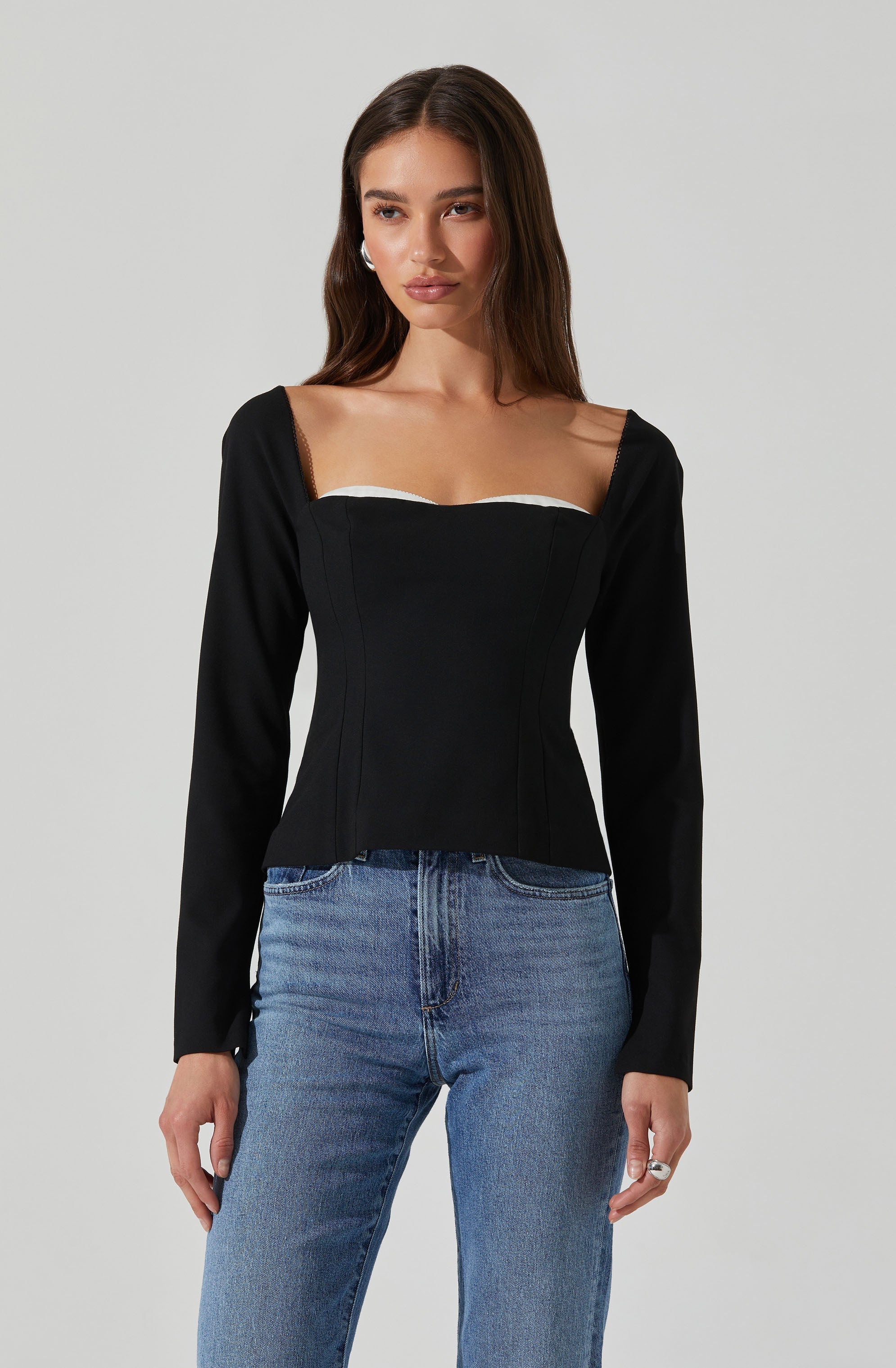 A woman is wearing a fitted black long-sleeve top with a square neckline that has a white trim detail along the top edge. She is also wearing light blue, high-waisted jeans.