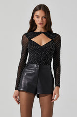 A woman is wearing a black, long-sleeved, sheer top with white polka dots and a distinctive cutout design at the chest. The top has a black bow detail at the neckline. She pairs it with high-waisted black leather shorts.