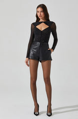 A woman is wearing a long-sleeved black sheer top with white polka dots and a cut-out detail at the chest area. She pairs it with high-waisted black leather shorts and black high heels with embellishments on the toes.
