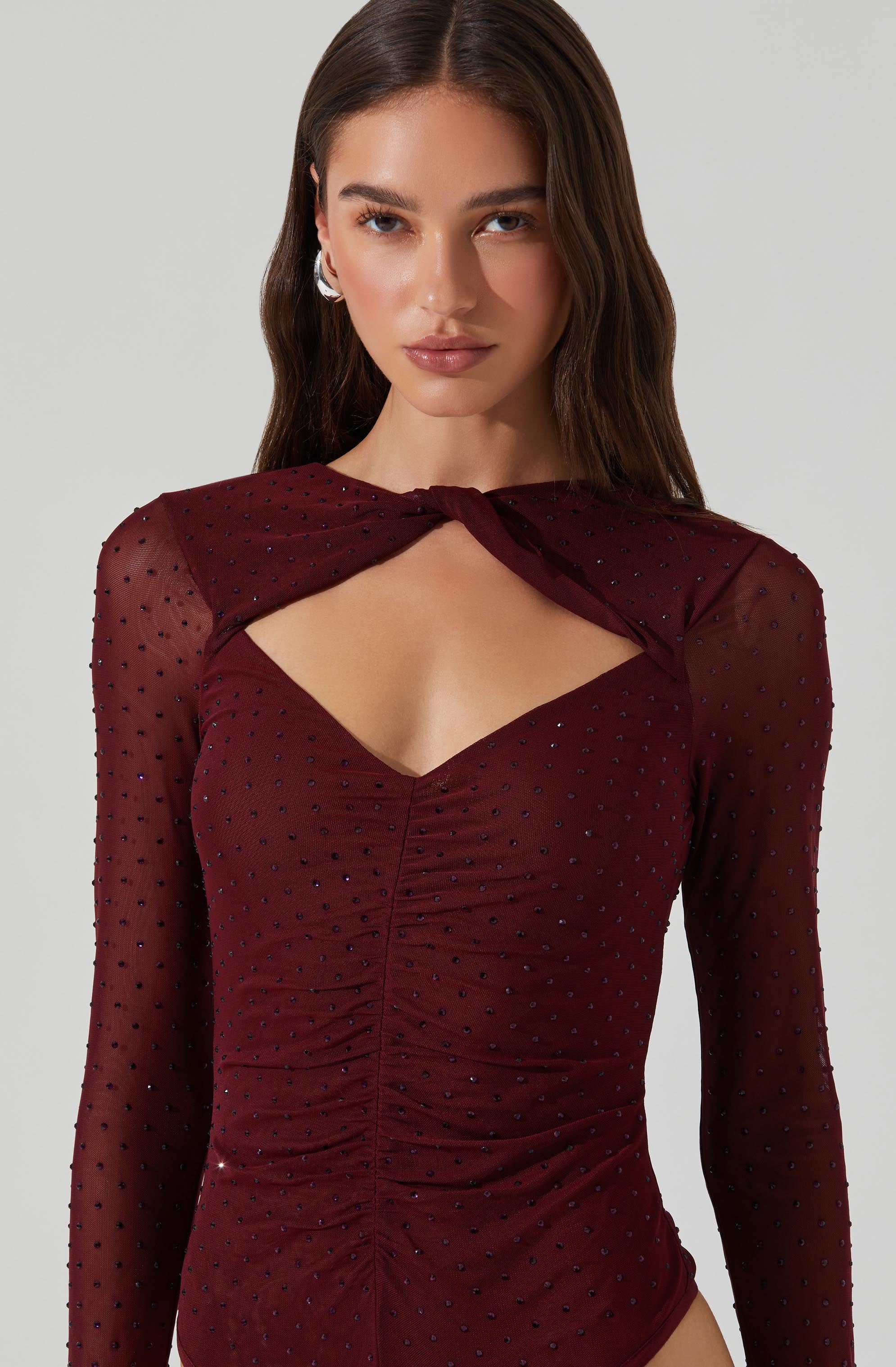 A woman is wearing a long-sleeved, deep red or maroon dress made of sheer fabric. The dress features small, shiny embellishments scattered throughout. It has a ruched front detail and an asymmetric, cutout neckline that reveals her collarbone and part of her chest. The dress also includes a matching, sheer cape-like overlay that drapes over her shoulders and connects at the front.
