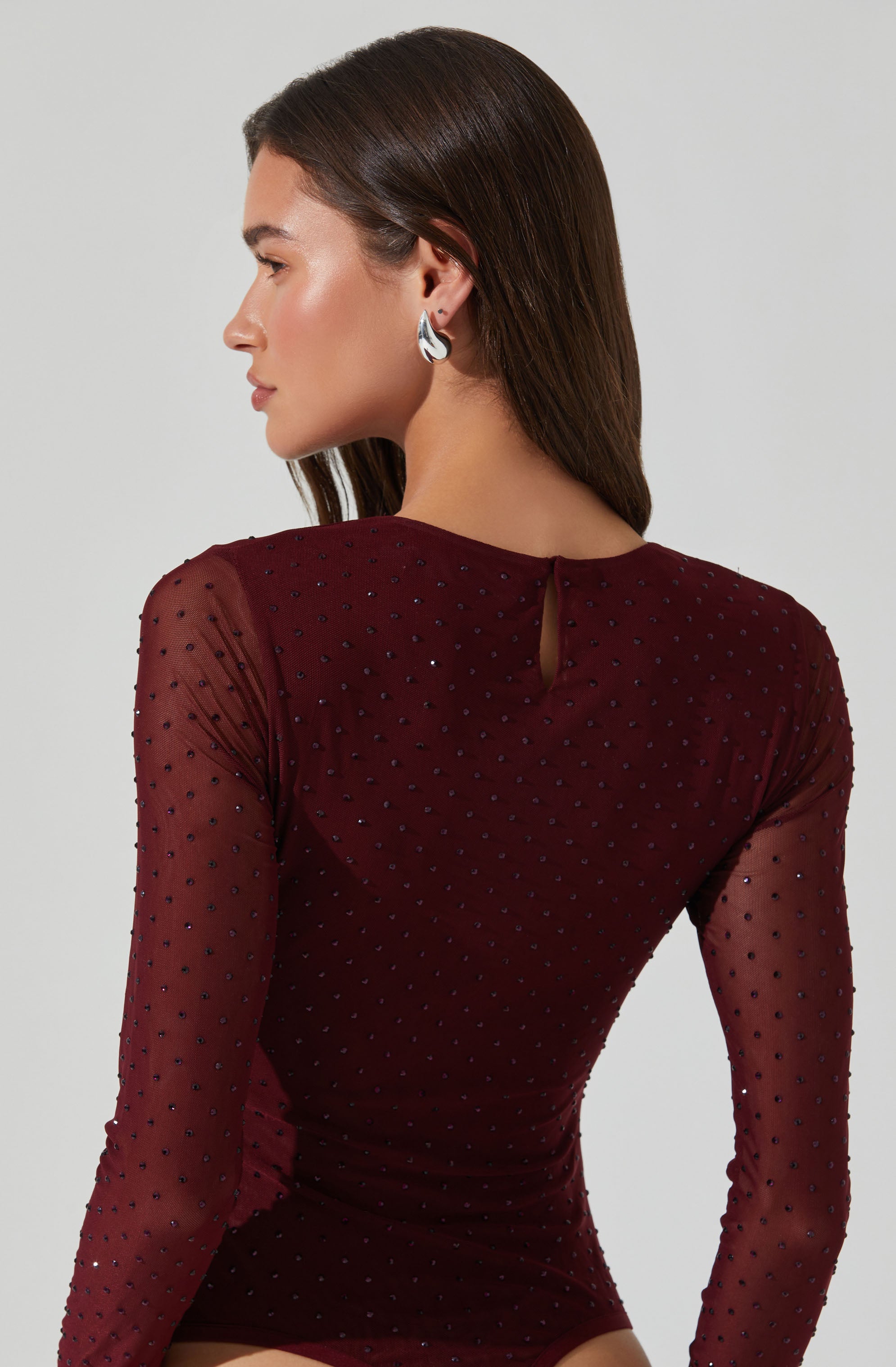 A woman is wearing a long-sleeved, form-fitting bodysuit made of sheer maroon fabric decorated with small, evenly spaced black dots. The bodysuit features a small keyhole opening at the back neckline.