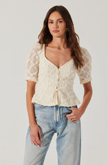 A woman is wearing an off-white, short-sleeved, lace blouse with a V neckline and a front tie detail. The blouse has a floral lace pattern and a slightly peplum silhouette. She pairs it with light-wash, high-waisted, loose-fitting jeans.