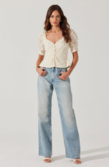 A woman is wearing a cream-colored lace top with short, puffed sleeves and a sweetheart neckline, featuring buttons down the front and a drawstring at the waist. She pairs it with high-waisted, light-wash, wide-leg jeans.