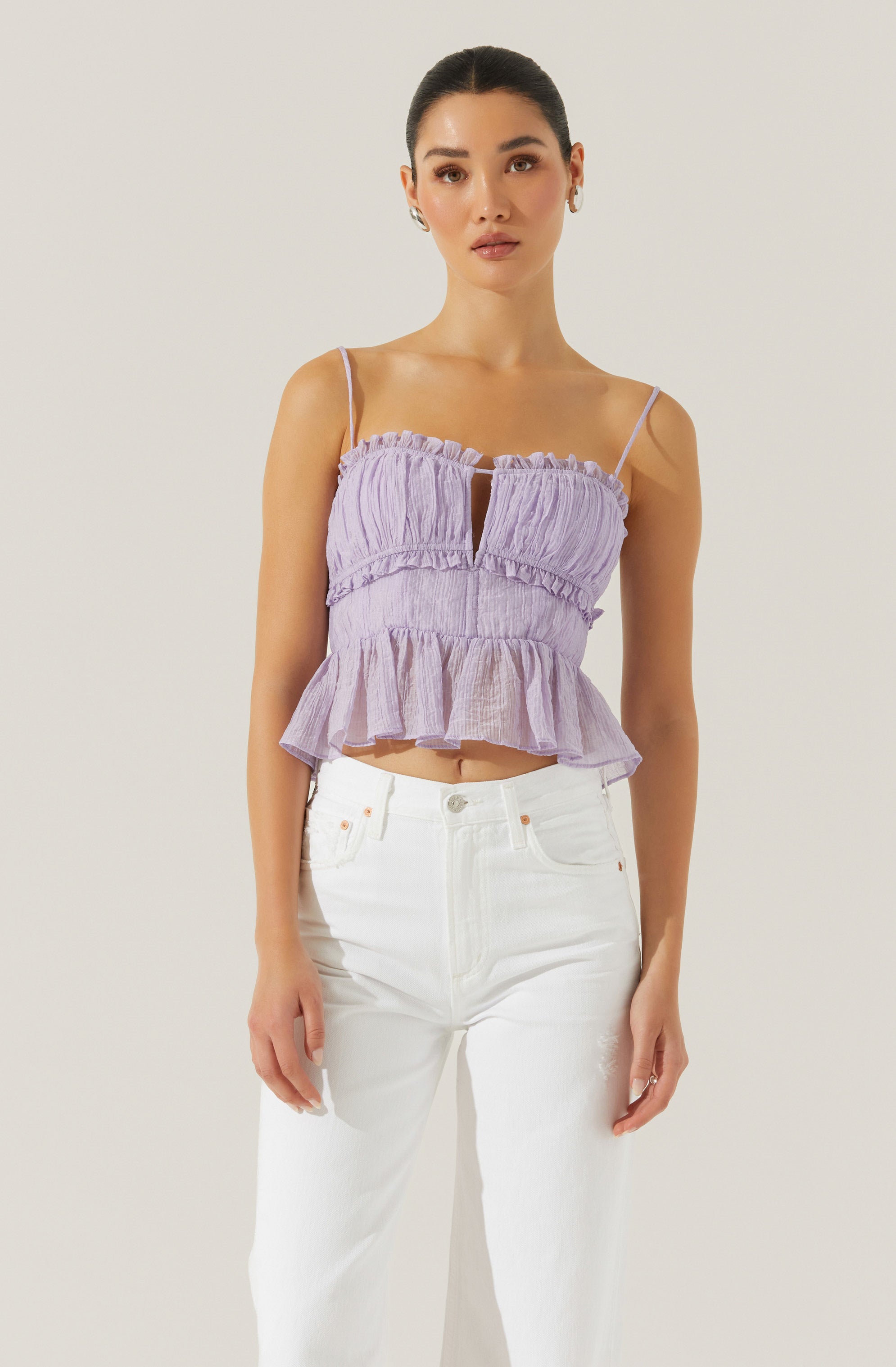 A woman is wearing a light purple, sleeveless, ruffled top with thin spaghetti straps and a keyhole opening at the front. She has paired it with high-waisted white jeans.