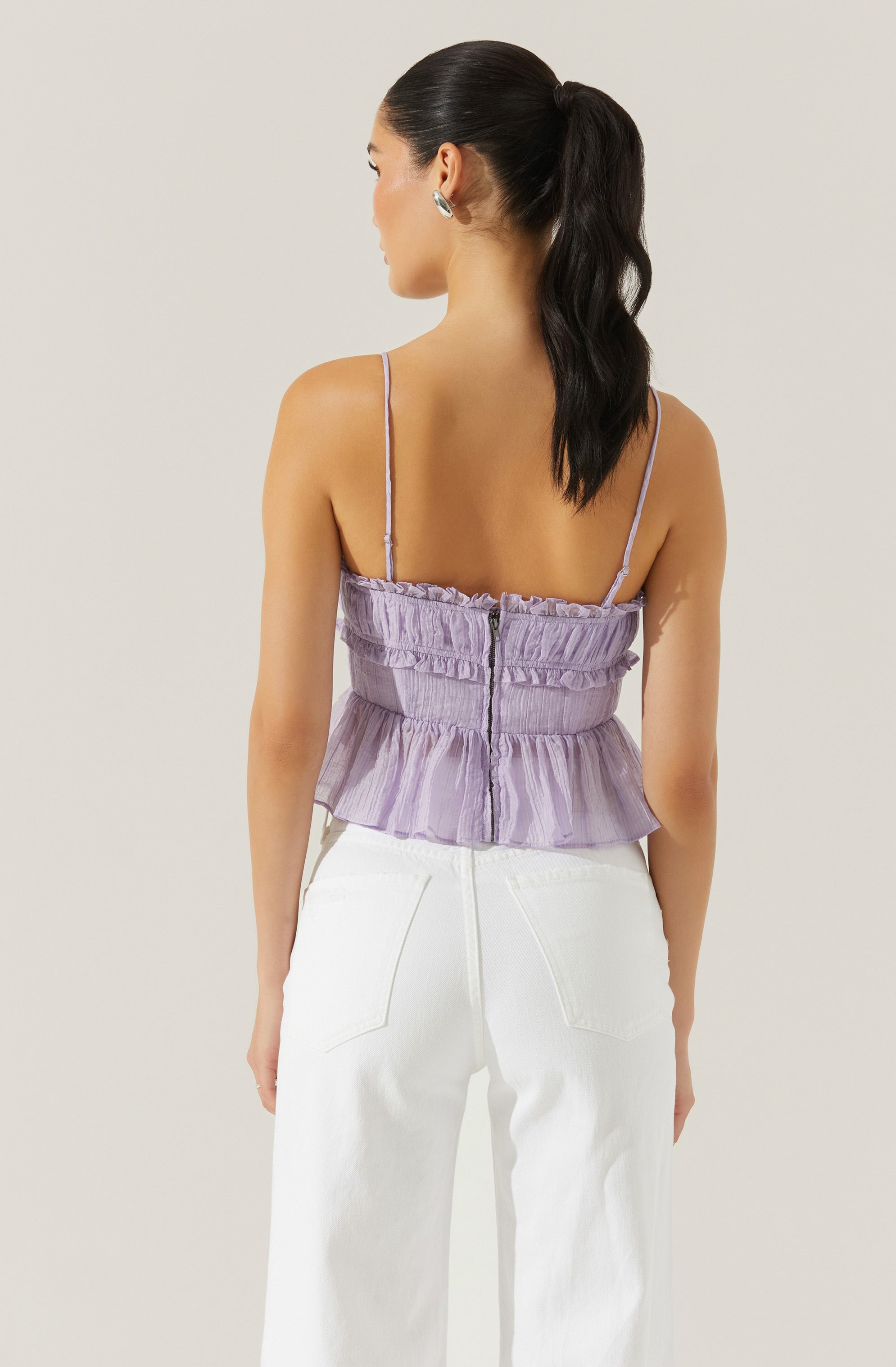 A woman is wearing a light purple, sleeveless top with thin spaghetti straps and ruffle detailing along the top and bottom edges. The top has a visible zipper closure at the back. She is also wearing high-waisted white pants with two back pockets.
