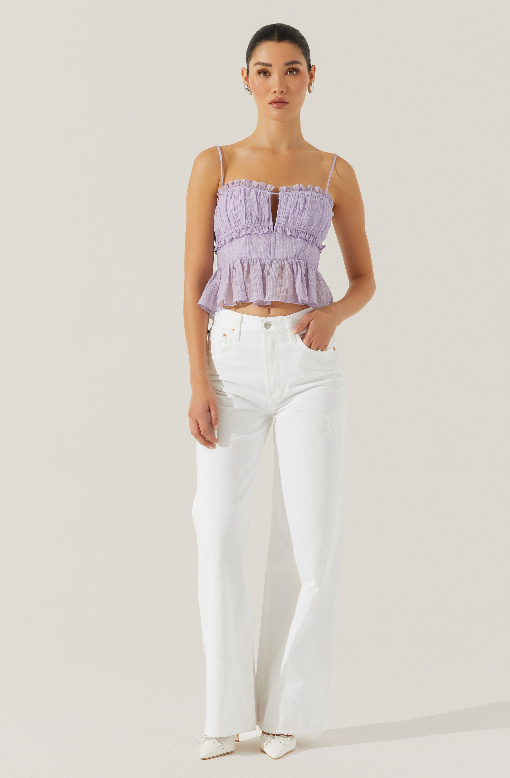 A woman is wearing a light purple sleeveless crop top with thin spaghetti straps and a keyhole opening in the center, featuring a ruffled hem. She pairs it with high-waisted white wide-leg jeans and white shoes.