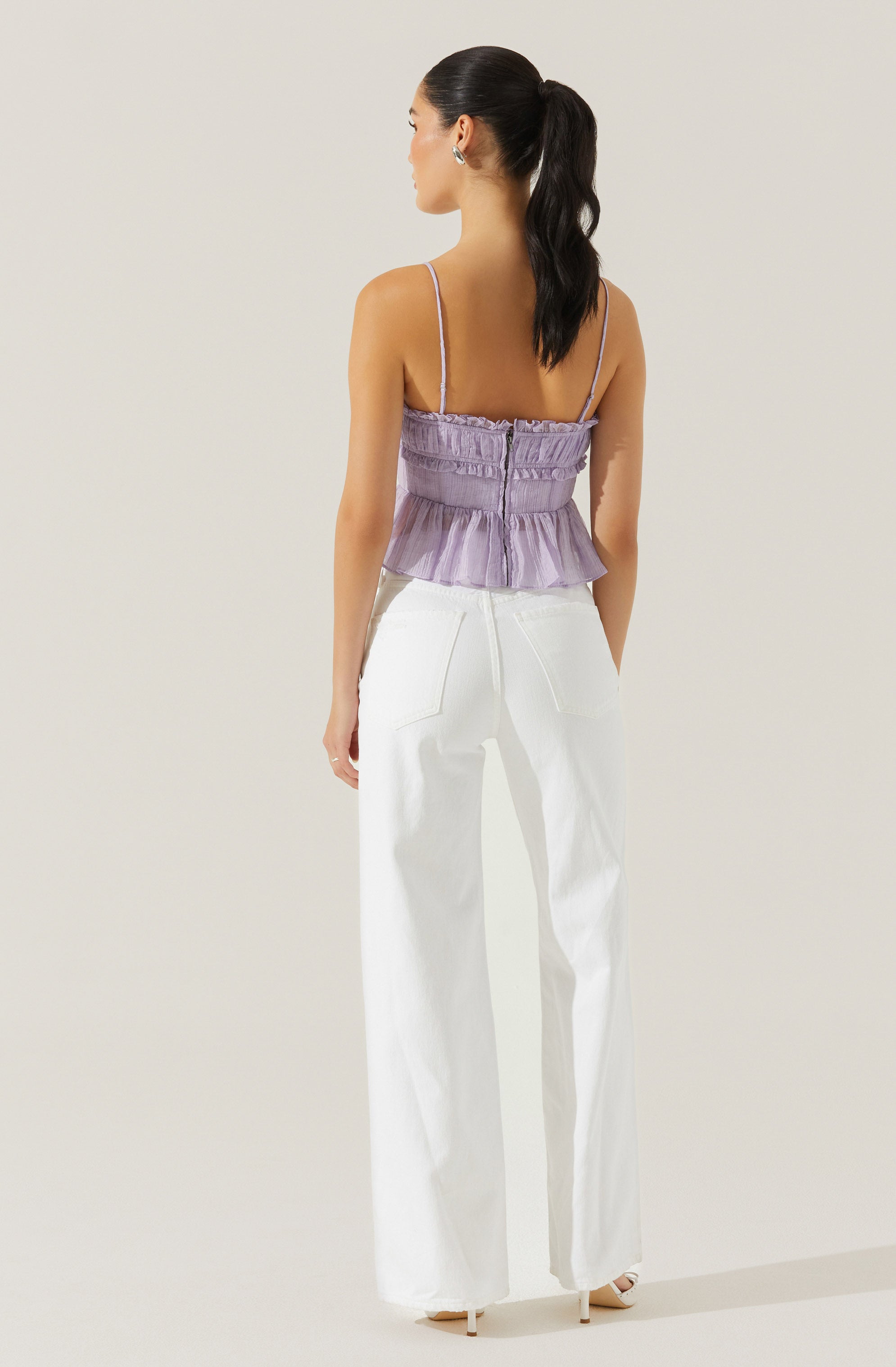 A woman is wearing a lavender, sleeveless, ruched crop top with thin spaghetti straps and a ruffled hem, paired with high-waisted, wide-leg white pants.