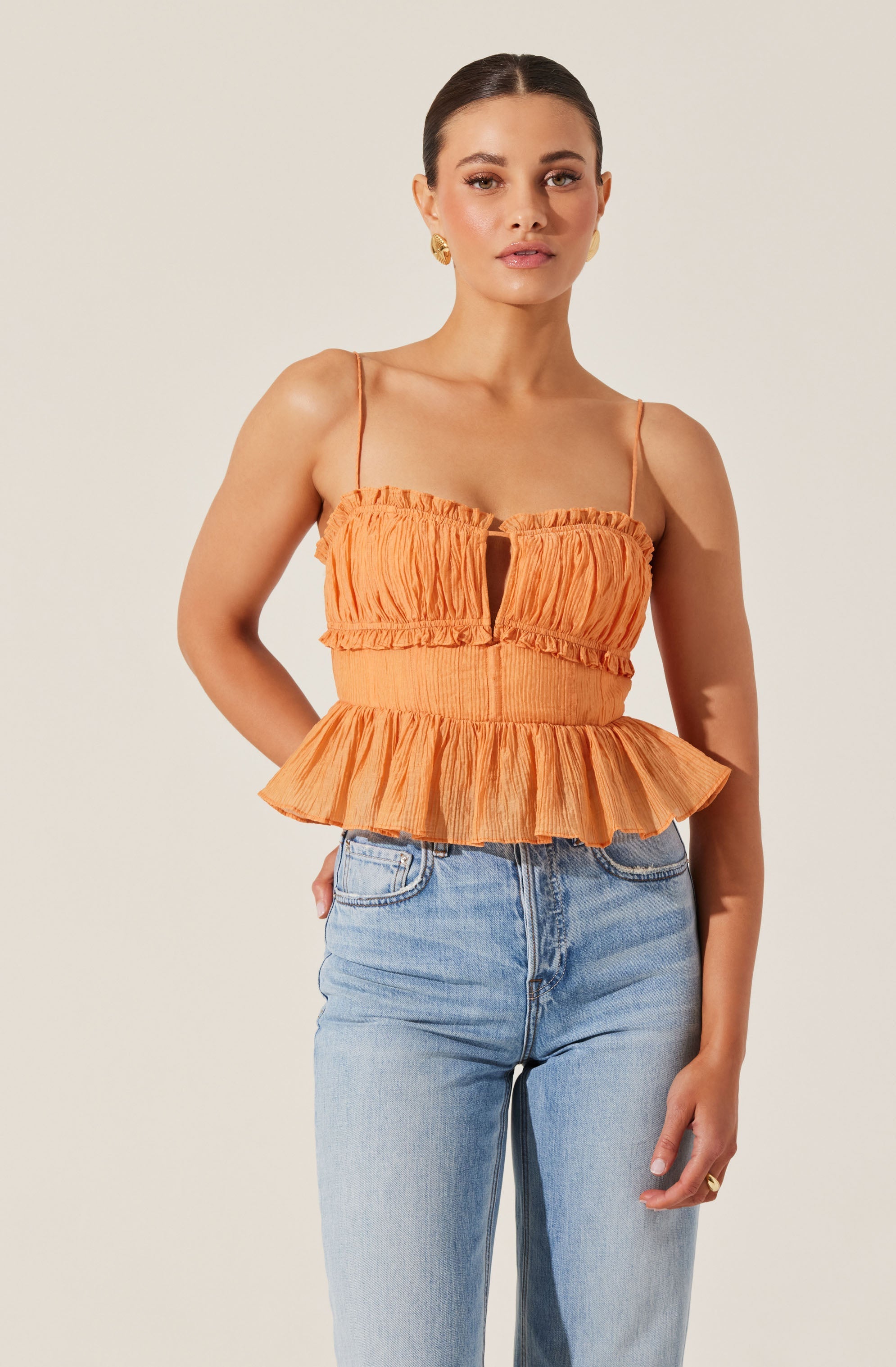 A woman is wearing an orange sleeveless top with thin spaghetti straps. The top features a gathered, crinkled fabric with a keyhole opening in the center of the chest. The waistline is elastic and ruffled, and the bottom of the top has a flared, ruffled peplum design.