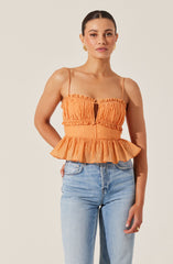 A woman is wearing an orange sleeveless top with thin spaghetti straps. The top features a gathered, crinkled fabric with a keyhole opening in the center of the chest. The waistline is elastic and ruffled, and the bottom of the top has a flared, ruffled peplum design.