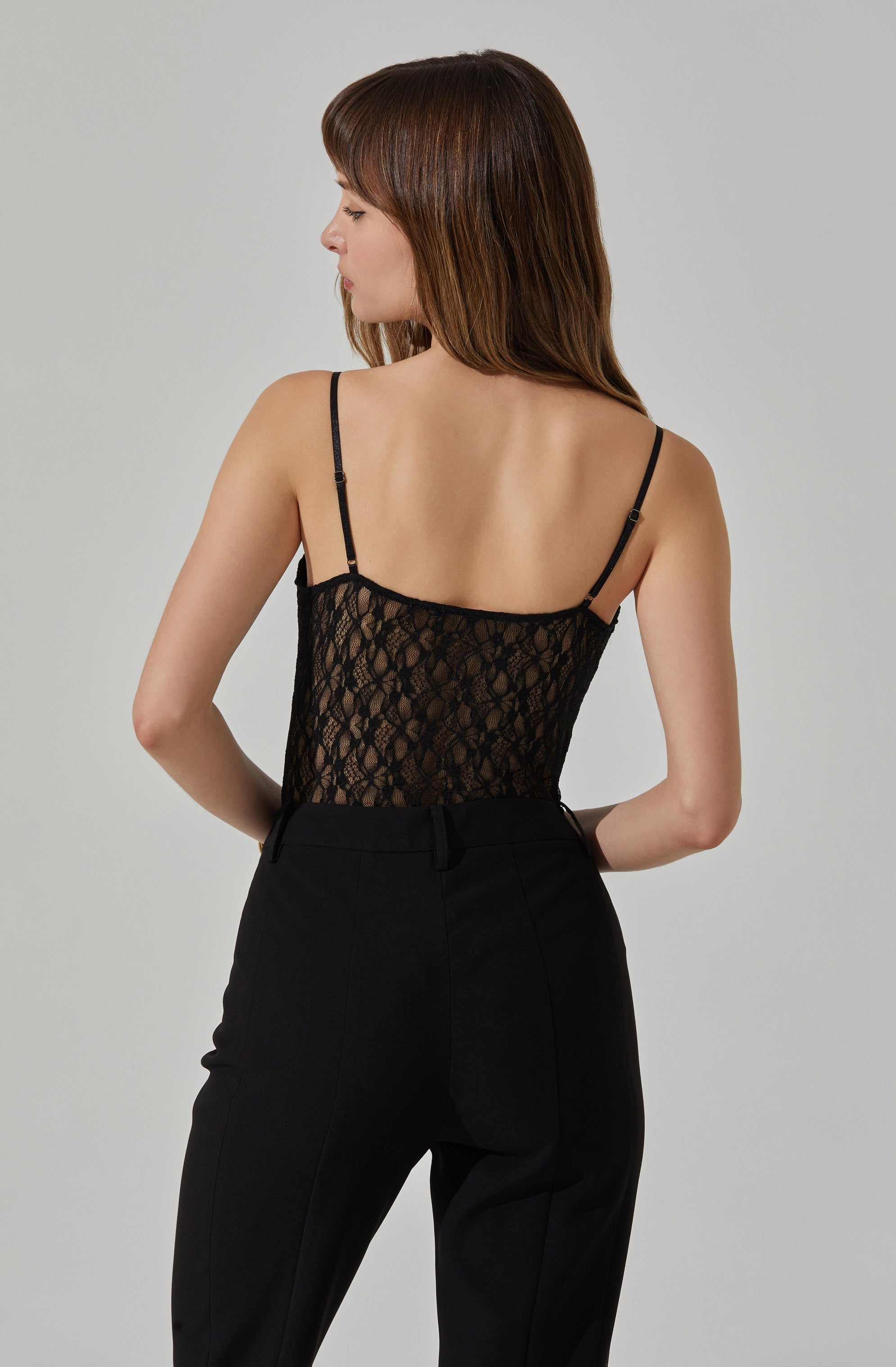 A woman is wearing a black sleeveless top with thin adjustable straps. The top is made of a lace fabric with a floral pattern. She pairs it with high-waisted black pants.