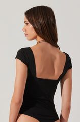 A woman is wearing a black short-sleeved top with a low, square back design. The top fits snugly and has a simple, elegant style.