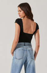 A woman is wearing a black top with short, slightly puffed sleeves and a wide, low back. She pairs the top with high-waisted, light-wash denim jeans that have prominent back pockets. Her brown hair is styled in loose waves, and she is turning slightly to the side, with her head turned towards her left.
