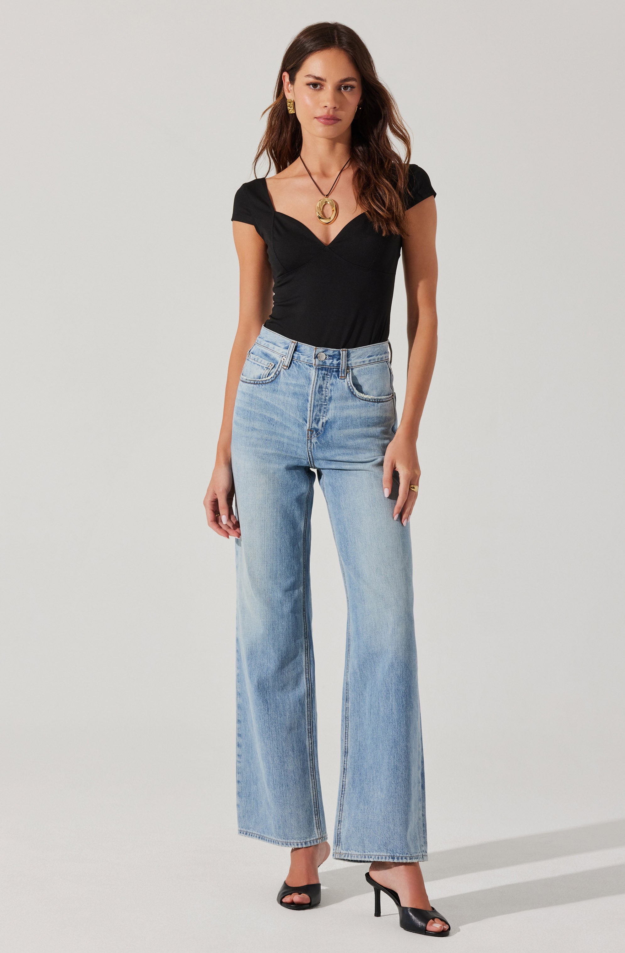 A woman is wearing a fitted black short-sleeve top with a sweetheart neckline, paired with high-waisted, light-wash, wide-leg jeans. She has on black open-toe high-heeled shoes, gold earrings, and a large gold pendant necklace.