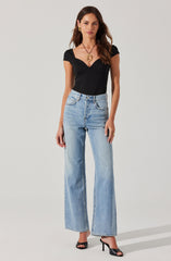 A woman is wearing a fitted black short-sleeve top with a sweetheart neckline, paired with high-waisted, light-wash, wide-leg jeans. She has on black open-toe high-heeled shoes, gold earrings, and a large gold pendant necklace.
