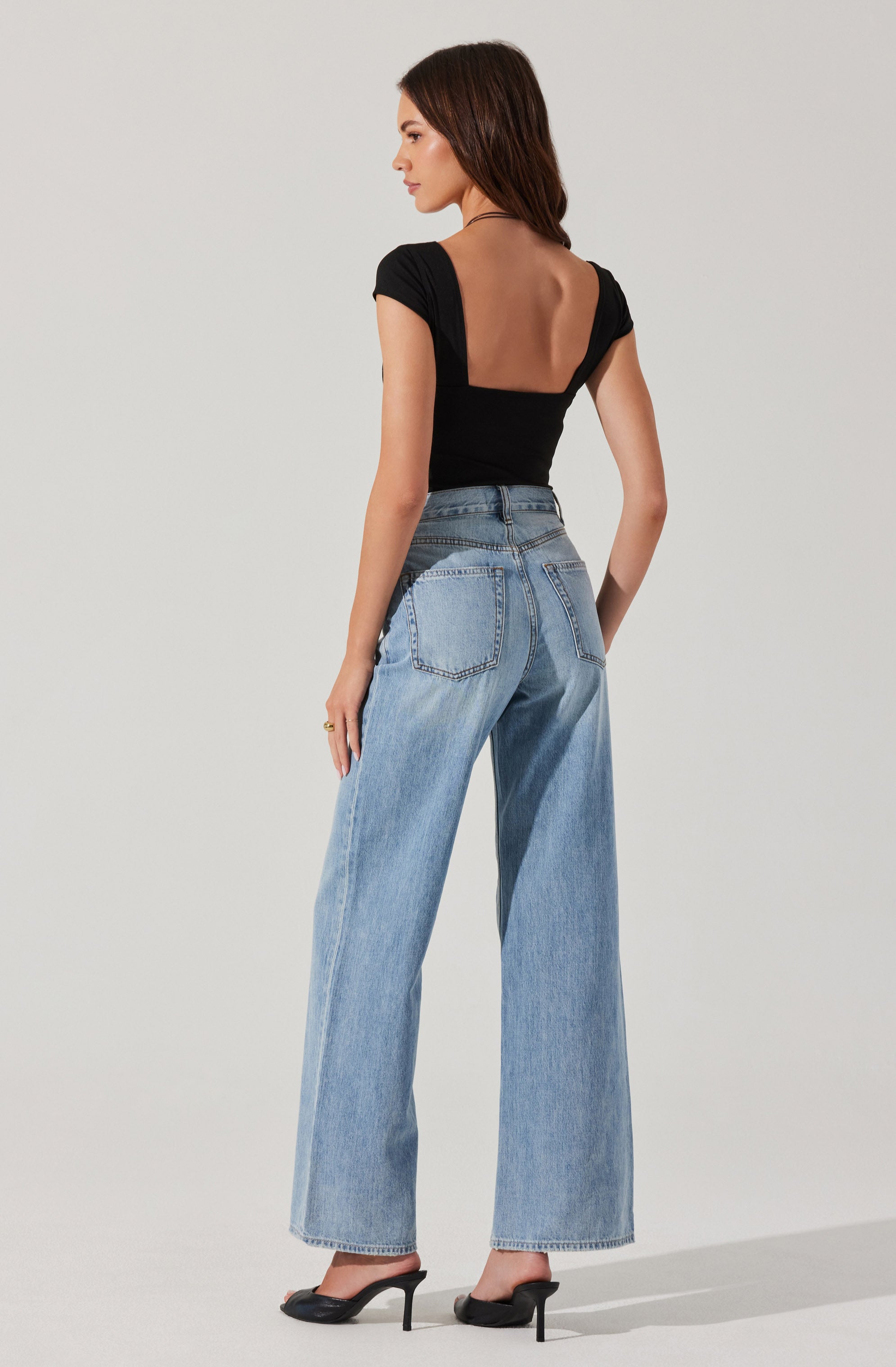 A woman is wearing a fitted black top with short sleeves and a scooped, exposed back. She is also wearing high-waisted, light blue, wide-leg jeans with visible back pockets. She has on black high-heeled shoes.