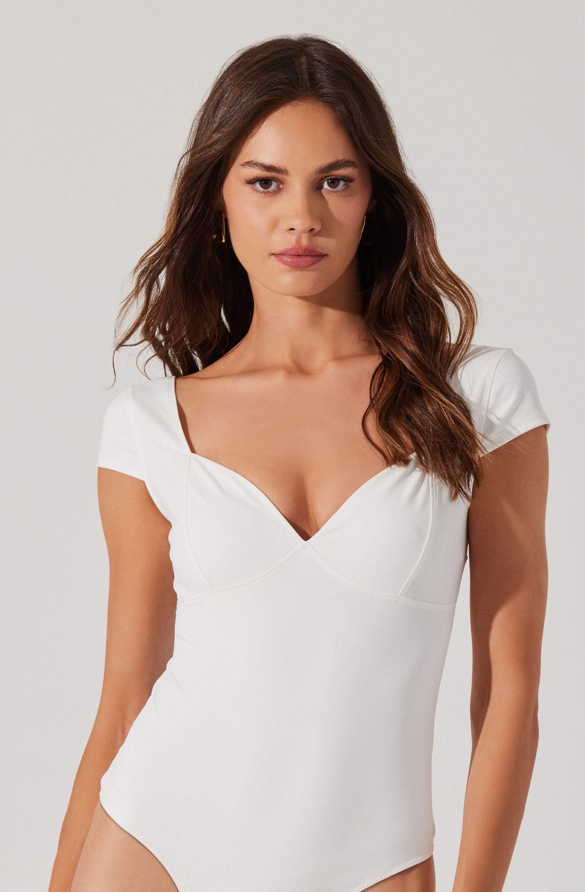 A woman is wearing a white, fitted dress with a sweetheart neckline and short sleeves. The dress has subtle seam details along the bust and waist, and the fabric appears smooth and slightly stretchy.