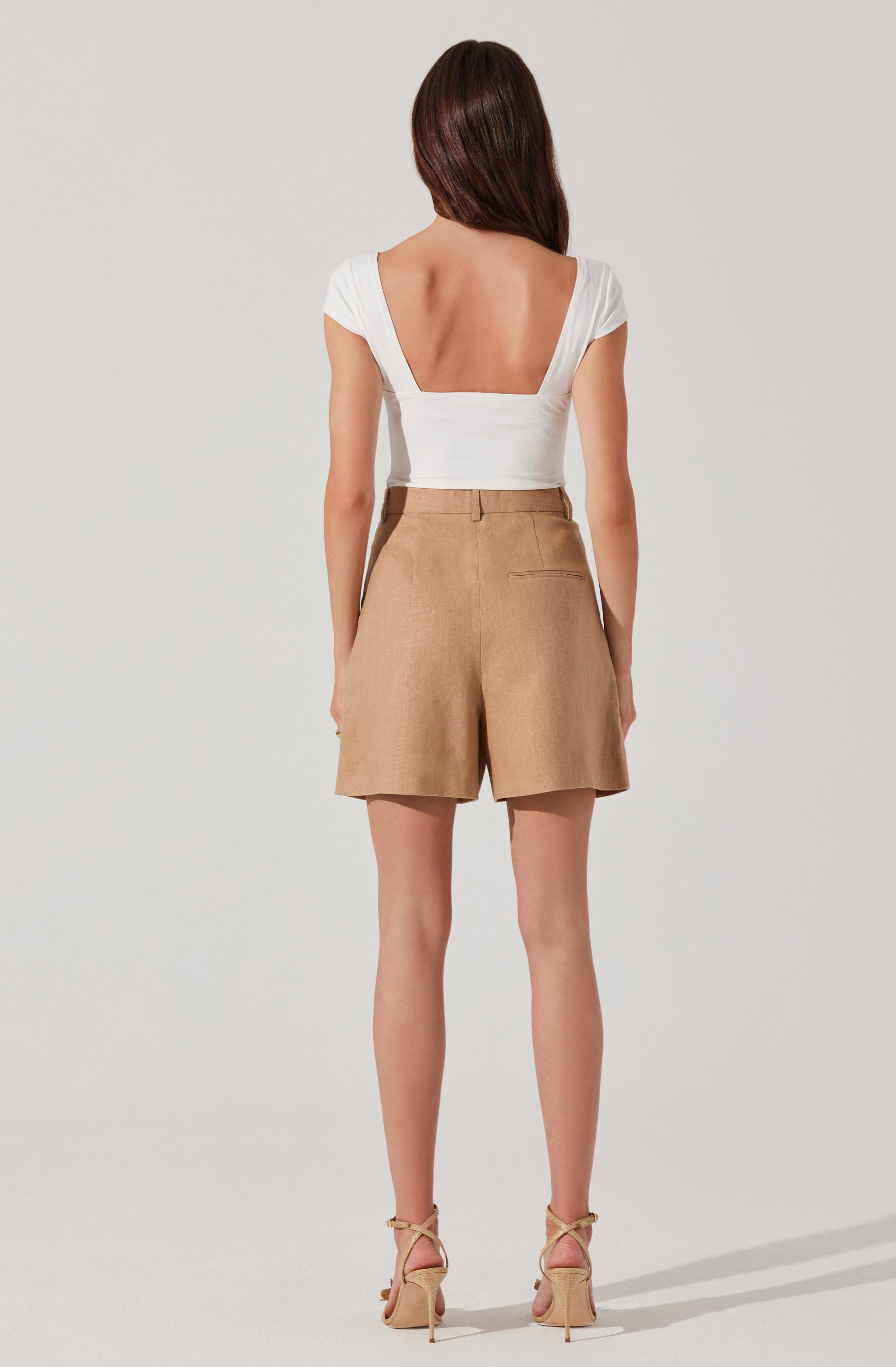 A woman is wearing a white, short-sleeved top with a deep, square neckline and a fitted waist. She pairs it with high-waisted, beige shorts that have a tailored fit and a single back pocket. She completes the outfit with nude, strappy high-heeled sandals.