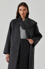 A woman is wearing a dark gray, oversized wool coat with a wide collar that drapes across her shoulder. Underneath, she has on a light gray, ribbed sweater. She is also wearing black high-waisted pants. She accessorizes with pearl stud earrings and a ring on her left hand.