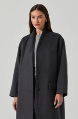 A woman is wearing a long, dark gray overcoat with a wide collar and a single button closure. The coat features a pocket on the front right side. The sleeves are slightly loose, and the overall style is sleek and minimalistic. She is also dressed in a light-colored top underneath.