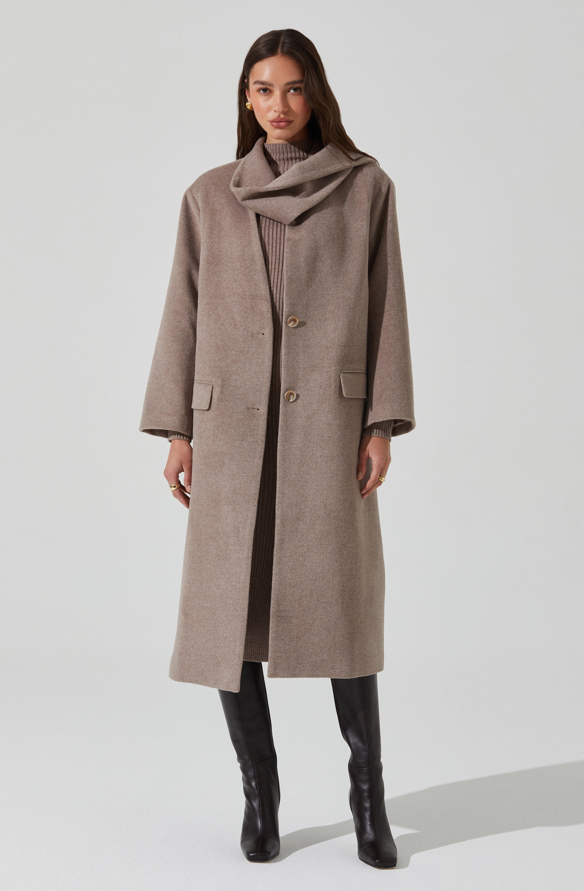 A woman is wearing a long, beige overcoat with a high collar and two large buttons near the waist. The coat features front pockets with flaps, and the sleeves are slightly loose. She pairs it with a matching beige turtleneck sweater underneath and black knee-high boots.