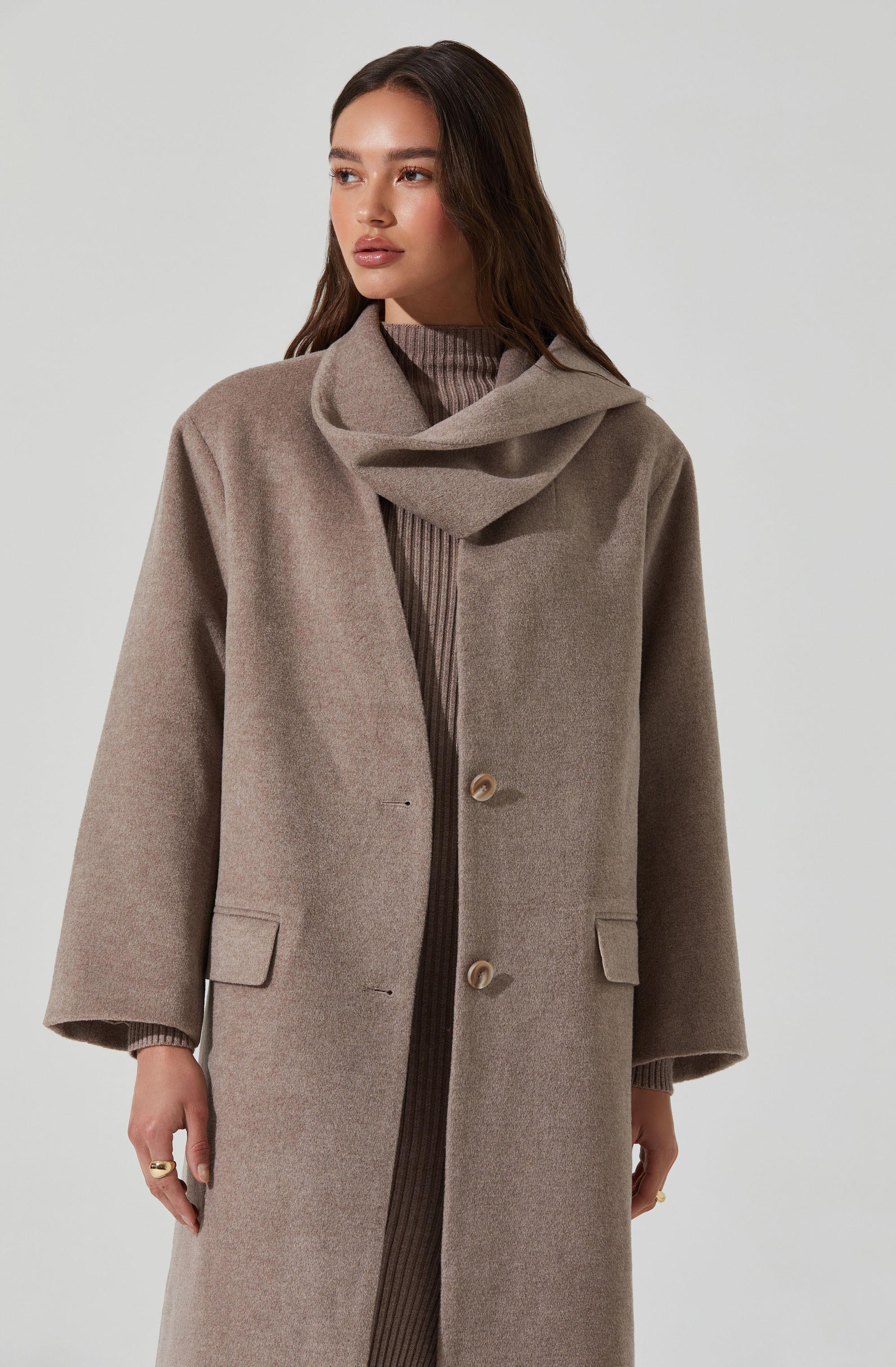 A woman is wearing a long, taupe-colored overcoat with a classic design, featuring two large front buttons and two flap pockets on either side. Underneath, she has on a ribbed, taupe-colored sweater with a high neckline. The coat has wide sleeves and a relaxed fit, complemented by a scarf or collar detail that drapes around her neck.