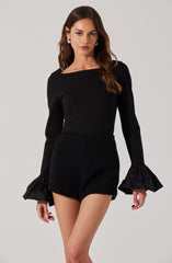 A woman is wearing a black long-sleeved top with a boat neckline and dramatic ruffled cuffs made of shiny fabric. She pairs it with high-waisted black textured shorts.