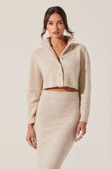 A woman is wearing a light beige, cropped, button-up cardigan with a high collar, paired with a matching high-waisted, fitted knit skirt.