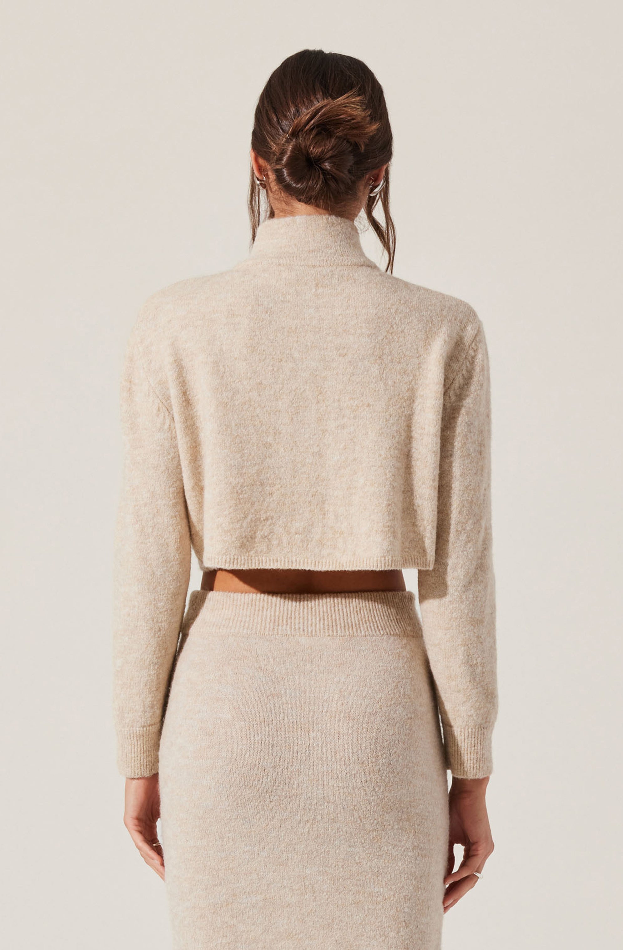 A woman is wearing a beige, cropped, long-sleeve sweater with a high neckline, paired with a matching high-waisted, fitted skirt. The sweater appears soft and slightly fuzzy, and the skirt is ribbed at the waistband. The woman's hair is styled in a loose bun with strands hanging down the sides.