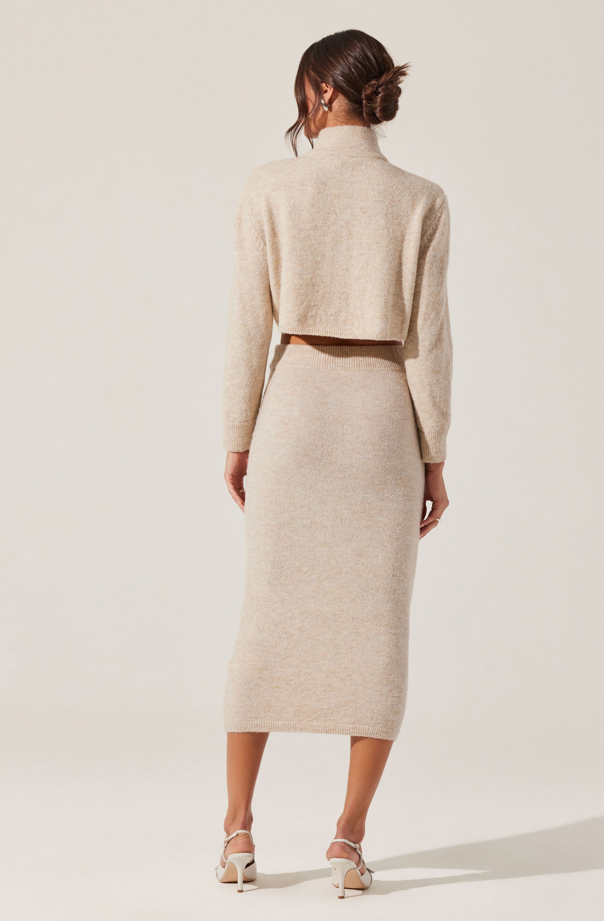 A woman is wearing a matching beige knit outfit consisting of a cropped, long-sleeved sweater with a high neckline and a high-waisted, midi-length skirt. She is also wearing white high-heeled sandals. The overall look is soft and cozy, with the textures of the knit fabric visible.