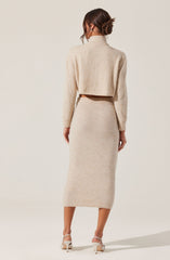 A woman is wearing a matching beige knit outfit consisting of a cropped, long-sleeved sweater with a high neckline and a high-waisted, midi-length skirt. She is also wearing white high-heeled sandals. The overall look is soft and cozy, with the textures of the knit fabric visible.