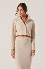 A woman is wearing a light beige, cropped, button-up sweater with a high collar and long sleeves. She is also dressed in a matching high-waisted, fitted, knit skirt that extends below the knees. The outfit appears cozy and has a soft, textured appearance.