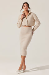 A woman is wearing a beige, cropped, zip-up fuzzy jacket with a high collar and a matching high-waisted, fitted pencil skirt that falls below the knees. She is also wearing a pair of beige pointed-toe heels with a bow detail on the front.