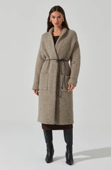 A woman is wearing a long, beige, textured coat made of a wool-like material, with a shawl collar and large front patch pockets. The coat is tied at the waist with a thin black drawstring, creating a cinched silhouette. Underneath, she has on a dark brown dress that reaches below the coat's hem. She completes her outfit with knee-high black leather boots that have a slight heel.