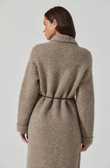 A woman is wearing a long, beige, textured knit coat with a high collar, and a thin black belt cinching the waist. The coat has a soft, slightly fuzzy appearance, with wide sleeves that are cuffed at the wrists.