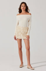 A woman is wearing an off-the-shoulder long-sleeve top in a cream color, paired with a cream-colored textured skirt that has a floral or ruffled pattern. She is also wearing cream-colored shoes with a strap detail.