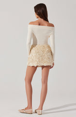 A woman is wearing an off-the-shoulder, long-sleeved white top with a fitted silhouette. She pairs it with a cream-colored, textured mini skirt featuring a floral or ruffled pattern. She is also wearing light-colored, patterned flat shoes.