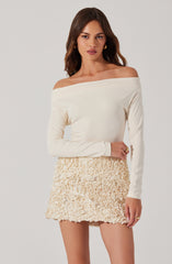A young woman is wearing an off-the-shoulder, long-sleeved, cream-colored top paired with a high-waisted, textured cream-colored miniskirt featuring a ruffled, floral-like pattern.