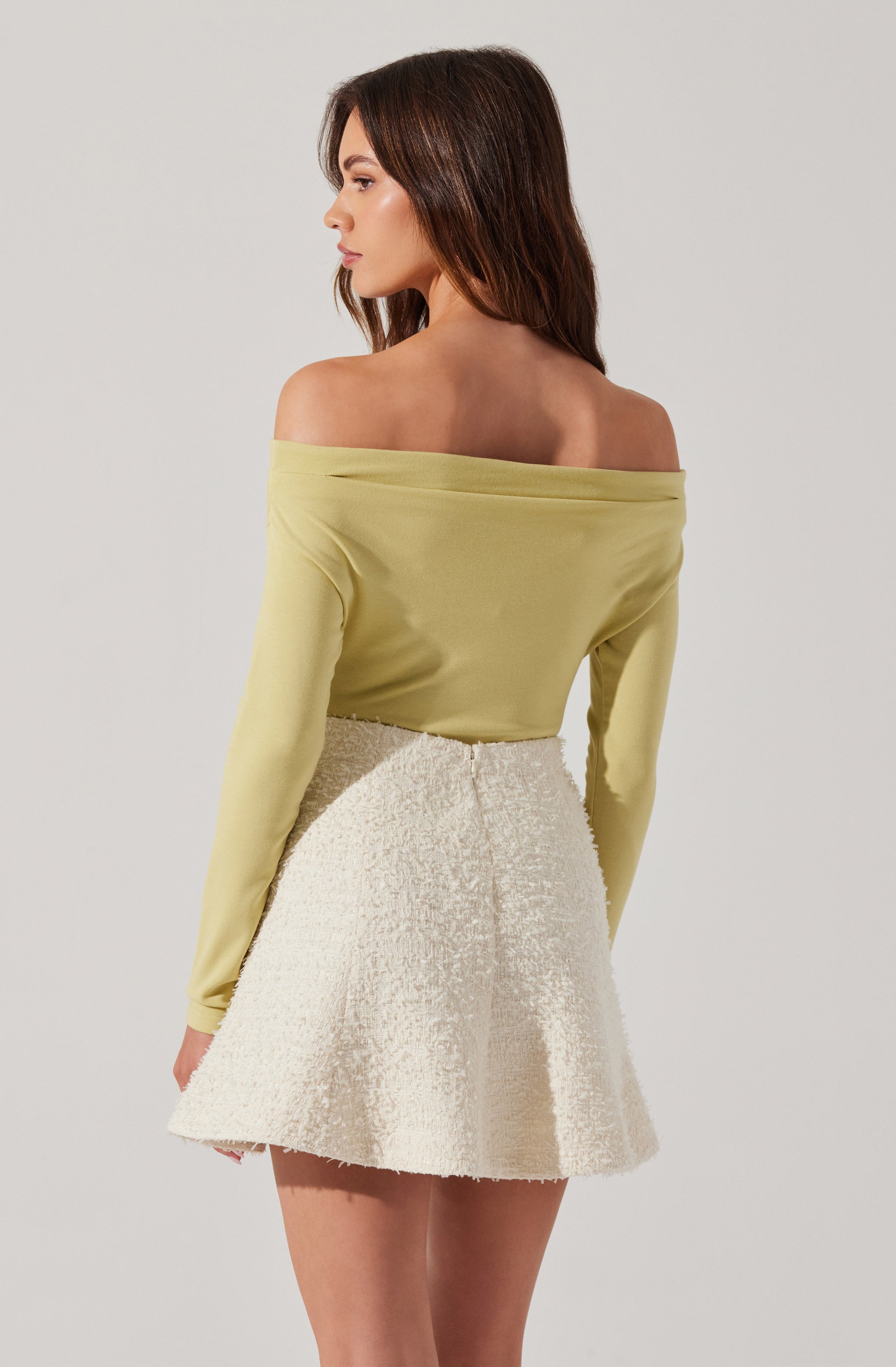 A woman is wearing an off-the-shoulder, long-sleeved, light yellow top paired with a high-waisted, cream-colored textured skirt that flares out slightly.