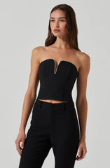 A young woman is wearing a strapless black crop top with a deep V neckline accented by a row of small, shiny embellishments along the edge. She pairs it with high-waisted black trousers.