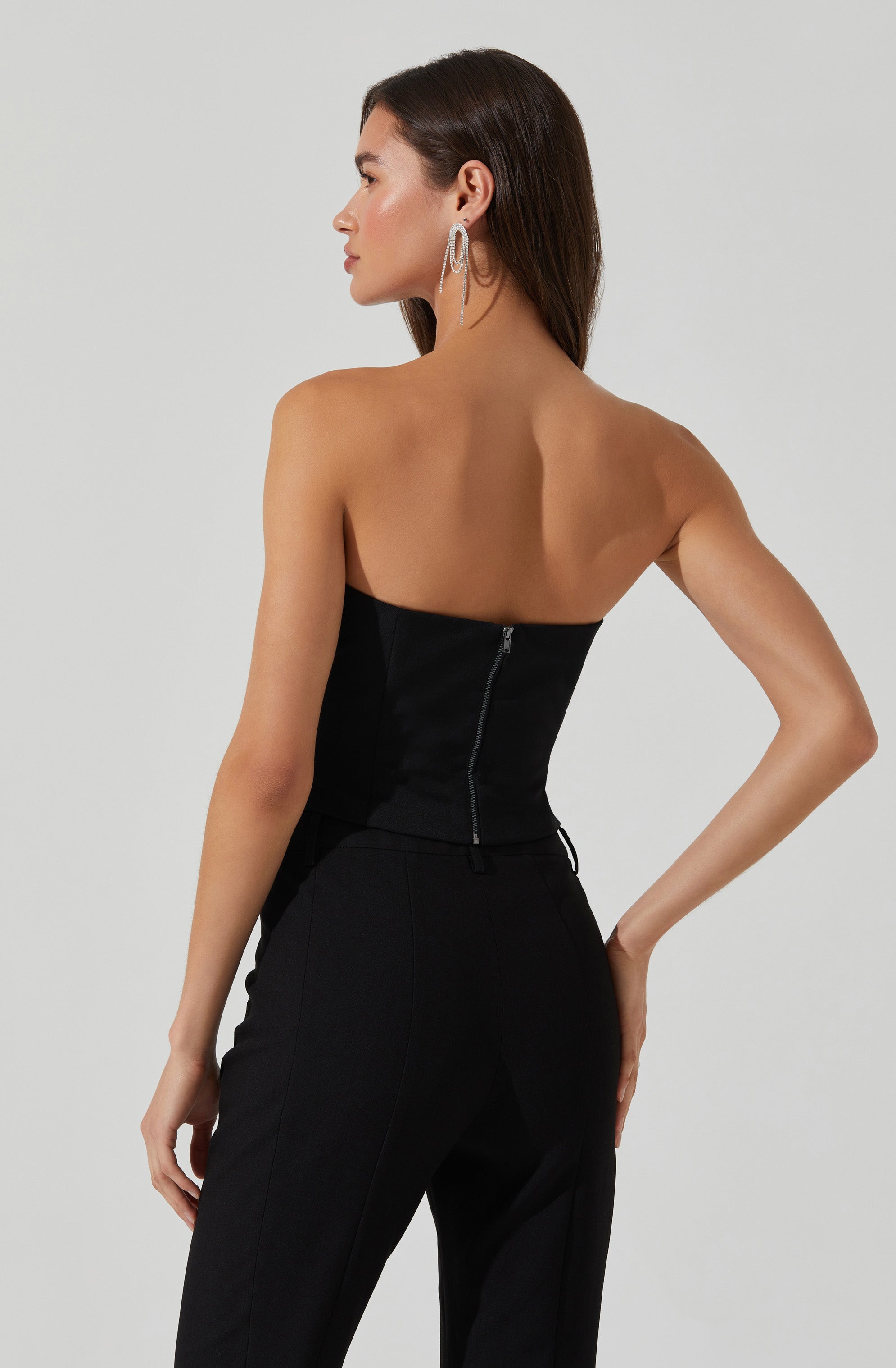 A woman is wearing a strapless black top with a visible zipper at the back. She pairs it with high-waisted black pants. She has long, dark hair and is accessorized with large, circular earrings.