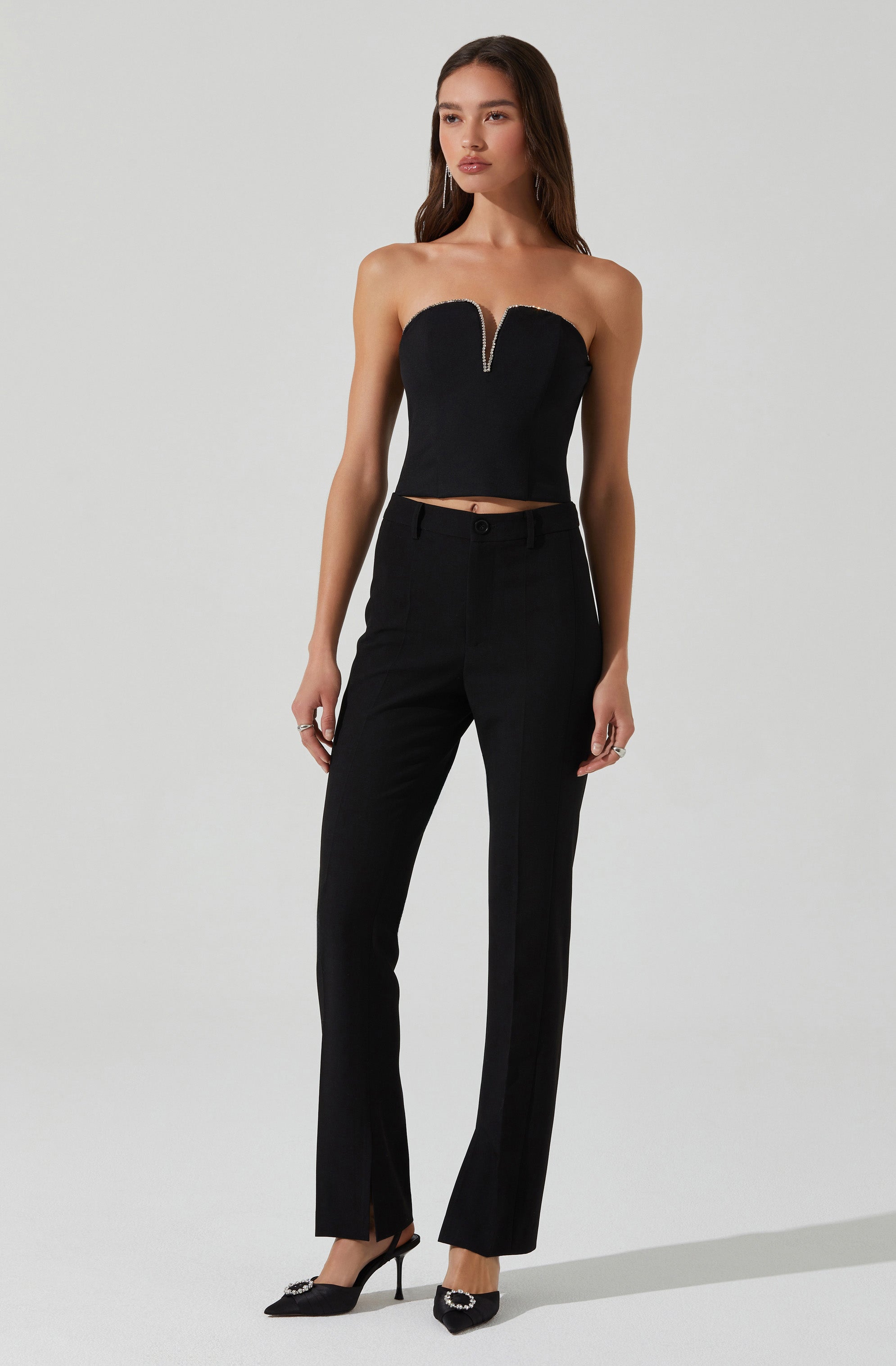A woman is wearing a strapless black crop top with a sweetheart neckline and a silver zipper down the front, paired with high-waisted black trousers that have a straight-leg fit and a small slit at the bottom of each pant leg. She is also wearing black pointed-toe heels with decorative embellishments.